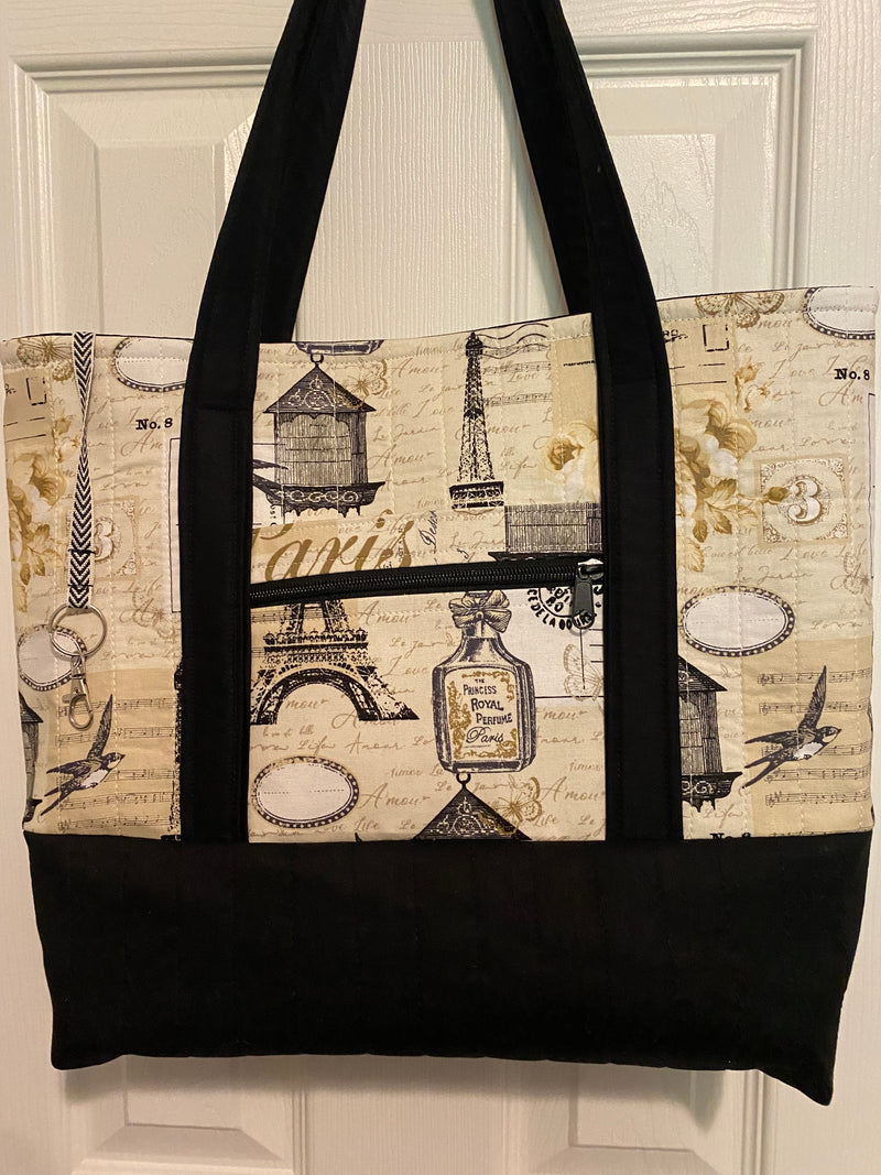 A tote bag with a vintage Paris-themed print featuring the Eiffel Tower, postcards, and script, with solid black straps and a solid black bottom.