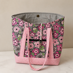 A fabric tote bag with a gray interior. Covered in a pink, black, and green floral pattern, featuring pink and white gingham check straps and a bottom base. Magnetic closure, two interior pockets, one exterior zippered pocket